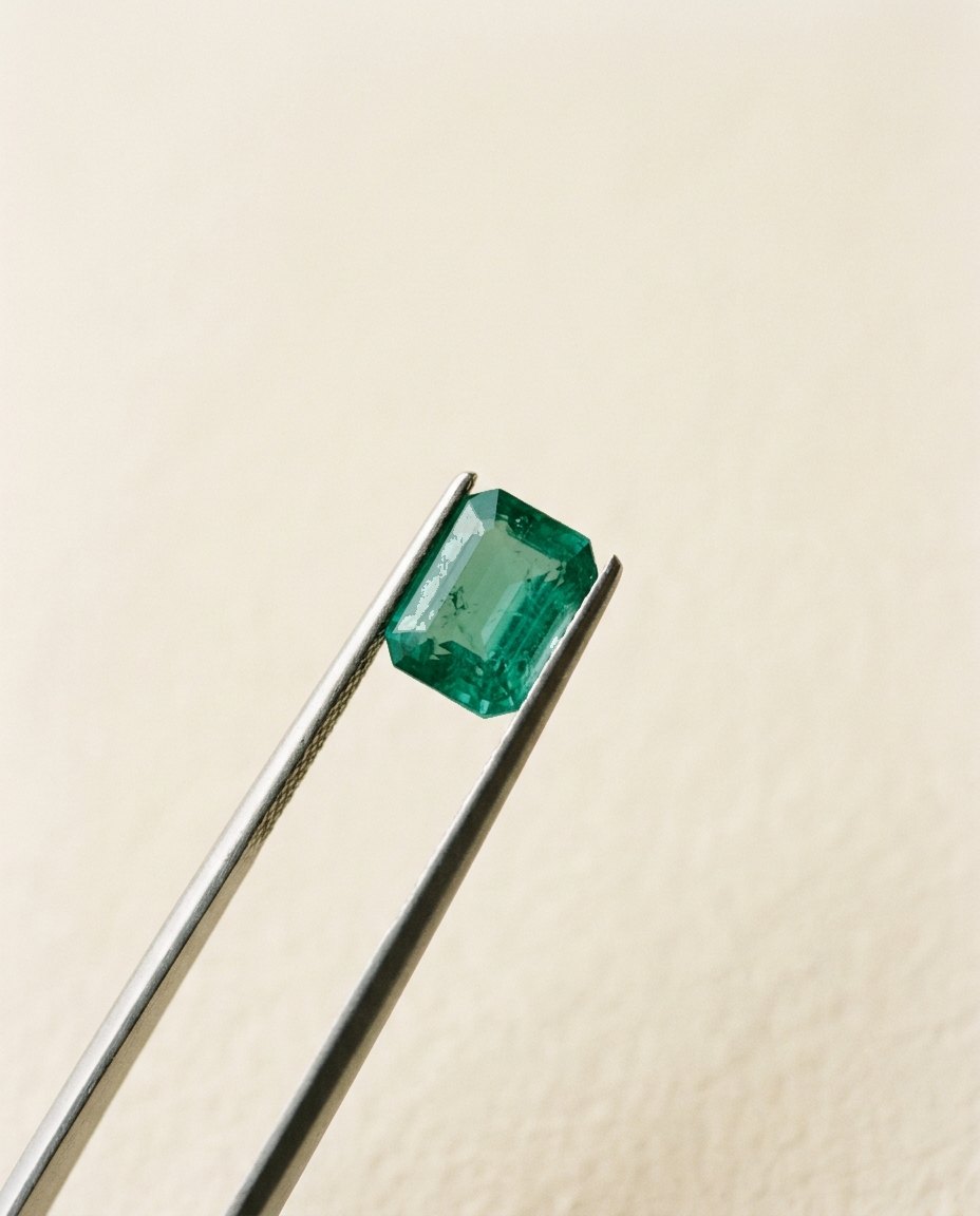 Steel gem tweezers holding a single emerald-cut emerald up to soft daylight